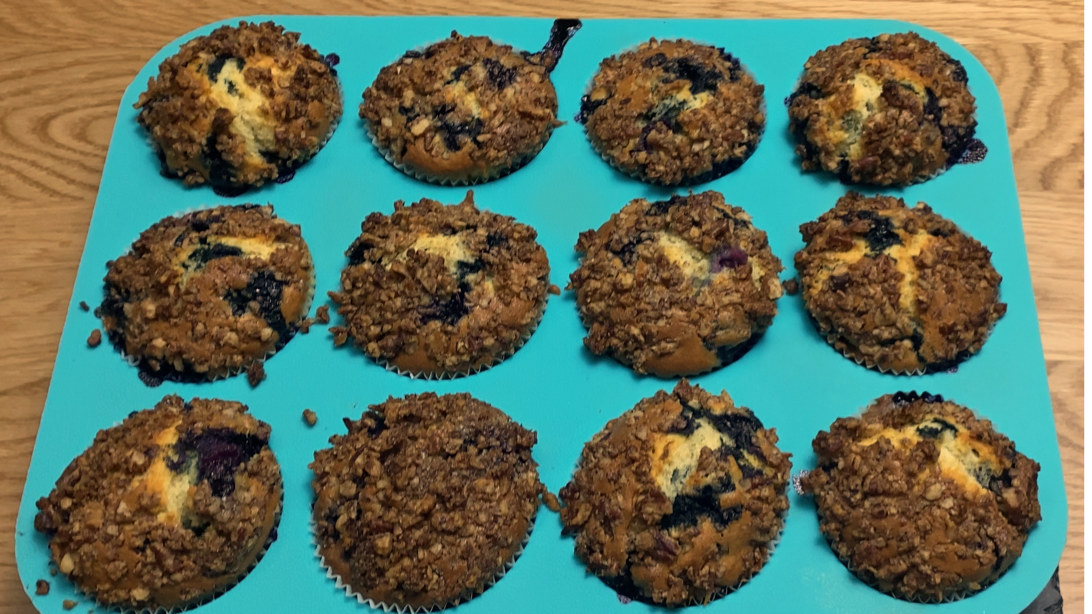 Twelve freshly baked blueberry muffins with a pecan streusel in a turquoise silicone muffin pan on a wooden surface.