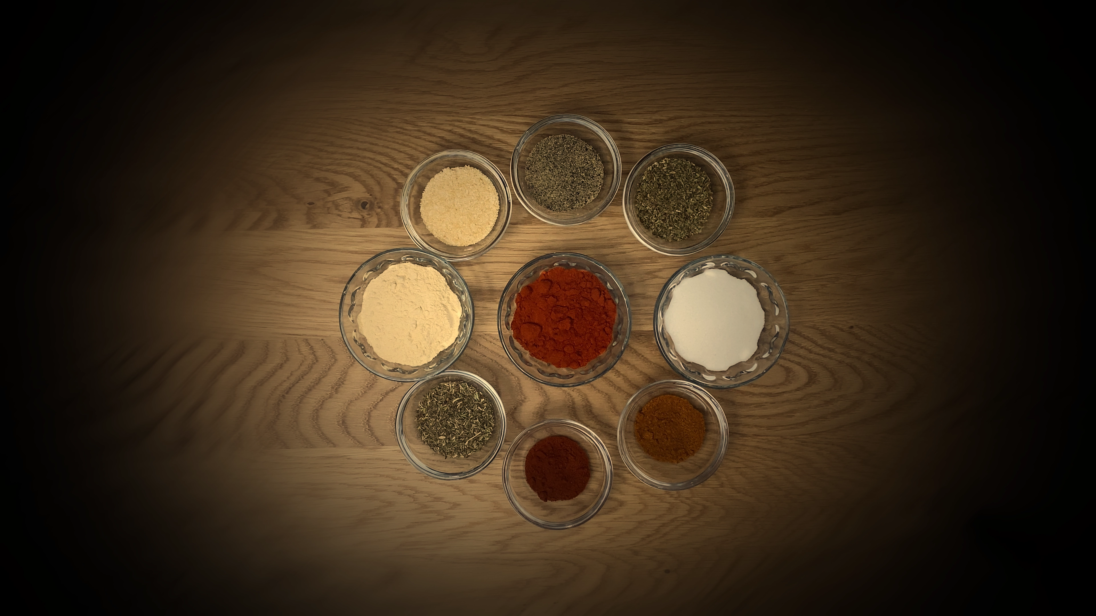 Top‑down photo of multiple clear glass bowls on a wood table, each filled with distinct Cajun spice ingredients like bright red paprika, salt, dried herbs, and assorted powdered seasonings for making homemade Cajun seasoning.