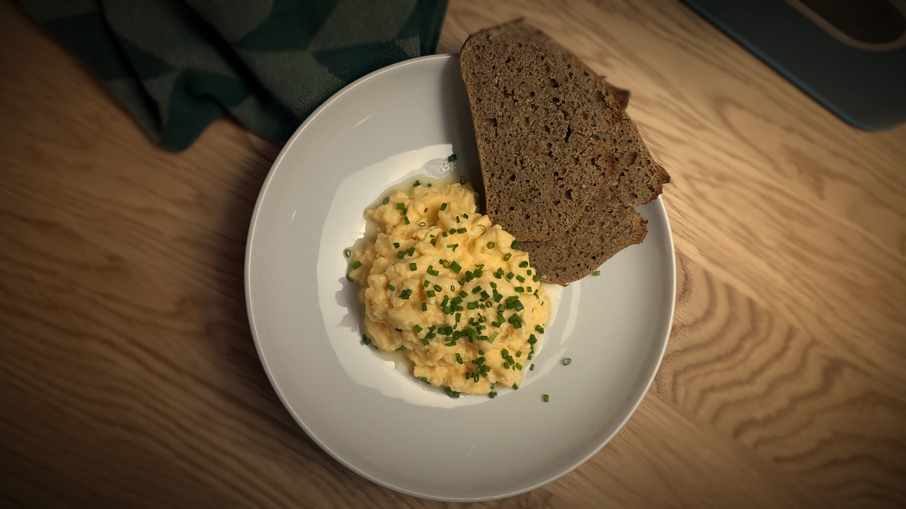 Top-down view of fluffy scrambled eggs topped with fresh green chives, served on a white plate with two slices of brown bread.