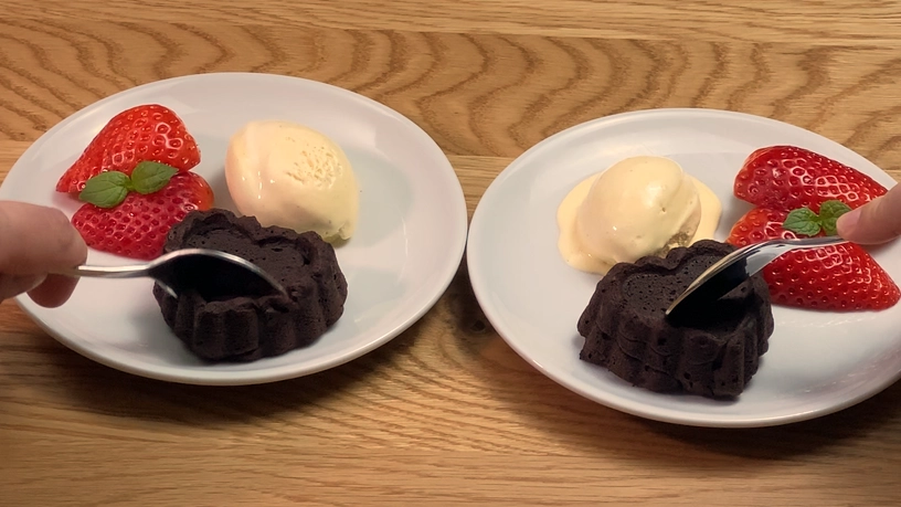 Two small dark chocolate brownies served on white plates with fresh strawberries and a scoop of vanilla ice cream on each plate. A hand holding a spoon is cutting into each brownie, all set on a wooden surface.