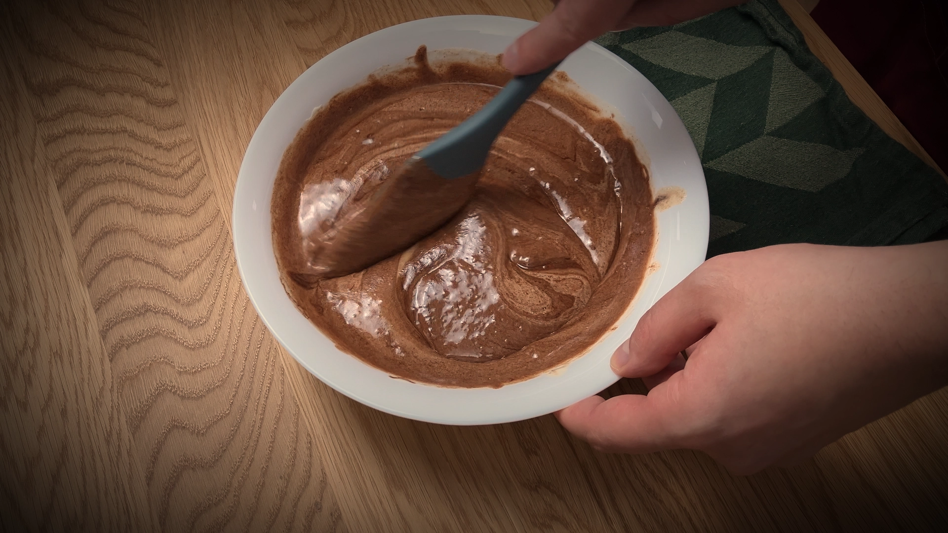 Hand stirring chocolate brownie batter in a white bowl with a gray spatula.