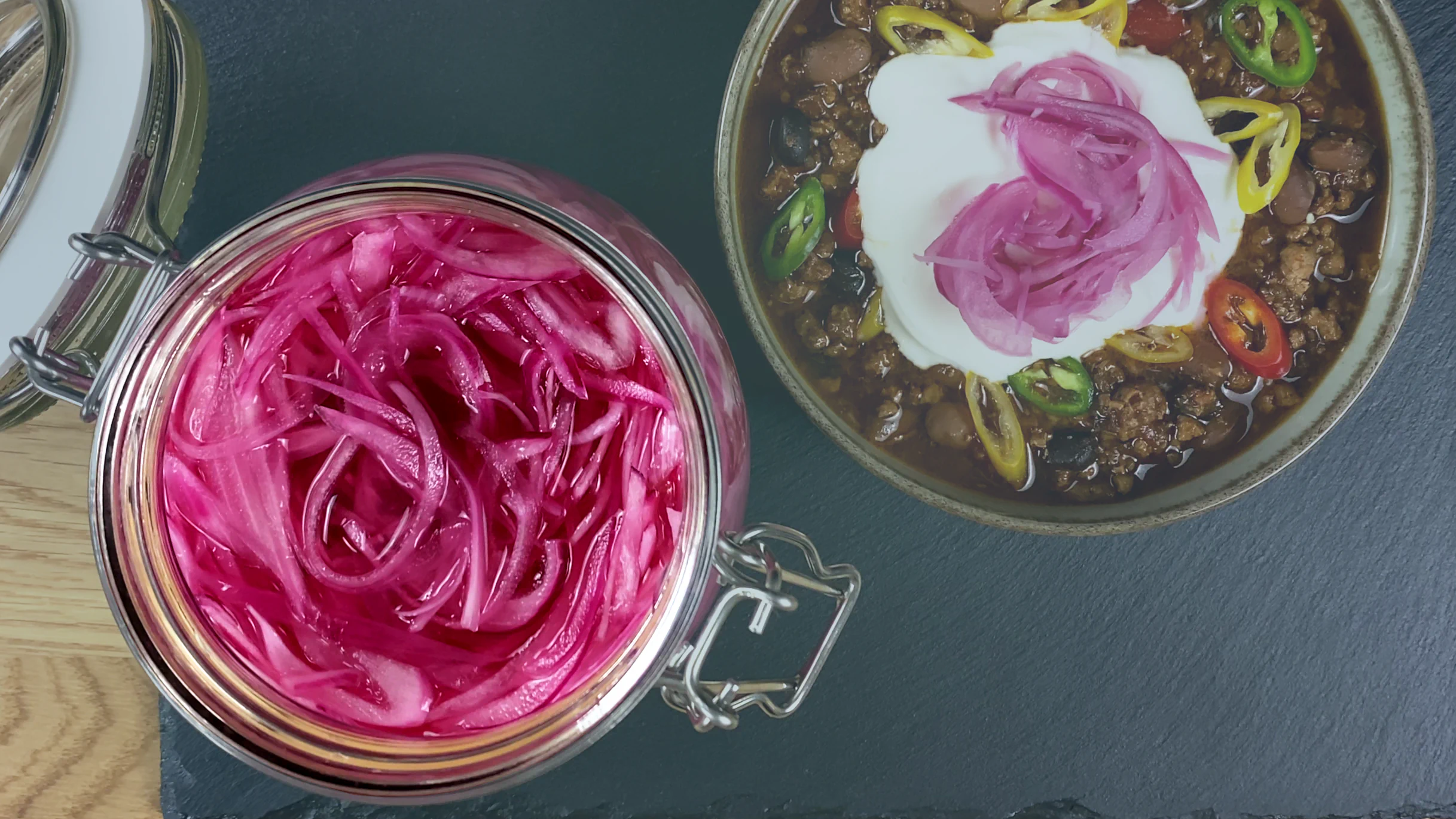 A top-down shot focusing on a jar of sliced pickled red onions. In the blurred background, a bowl of chili con carne is visible, topped with a dollop of Greek yogurt, fresh chilies, and pickled peppers.