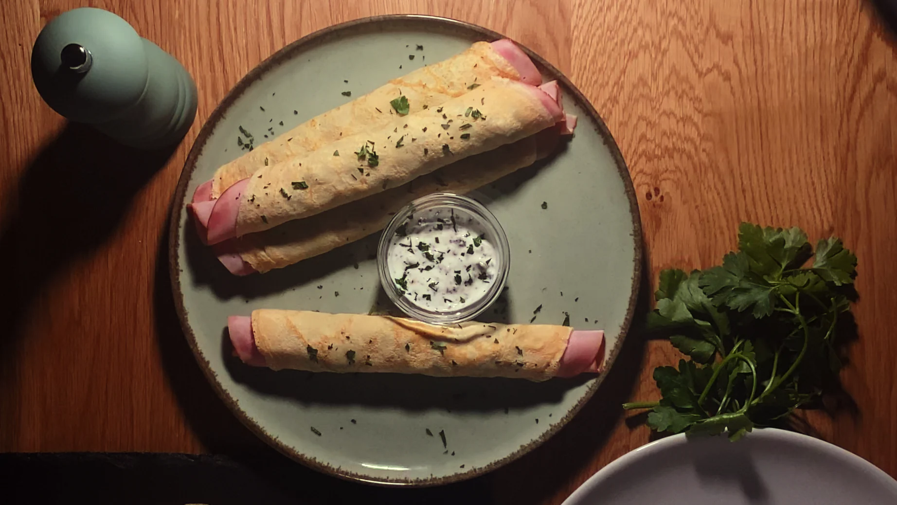 Top-down photo of a plate with a small bowl of mushroom sauce separating three ham crepes from a fourth. Accompanied by a pepper cracker and a bunch of fresh parsley.
