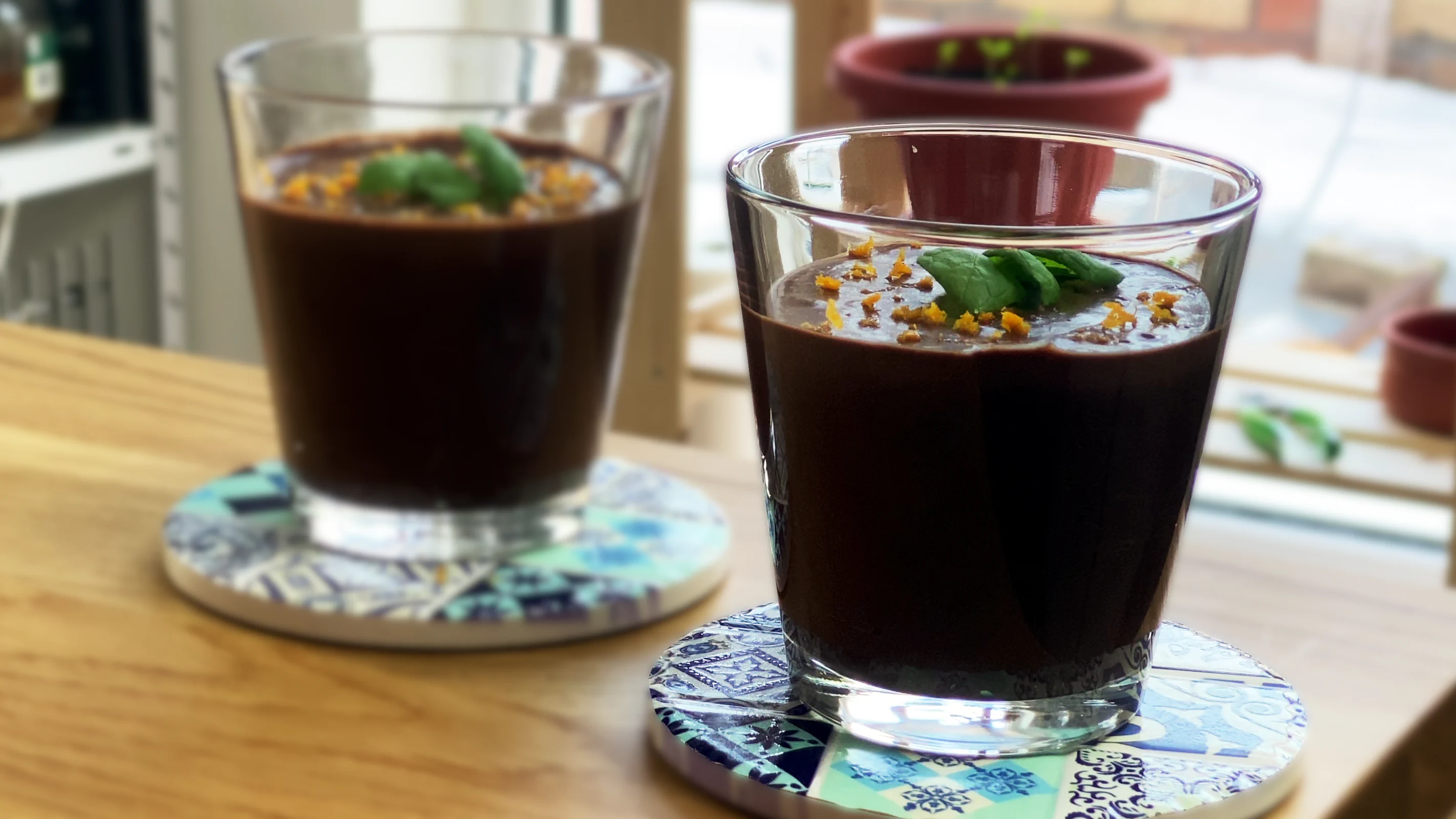 Two glasses of vegan chocolate mousse topped with fresh mint leaves and orange zest, served on decorative coasters by a sunlit window.